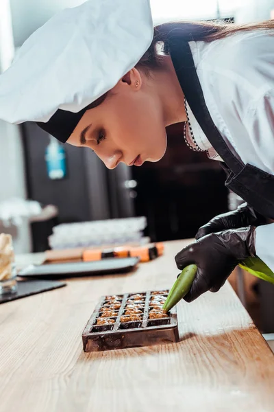 Vue latérale du chocolatier dans des gants en latex tenant un sac à pâtisserie avec des noix caramélisées près des moules à chocolat — Photo de stock