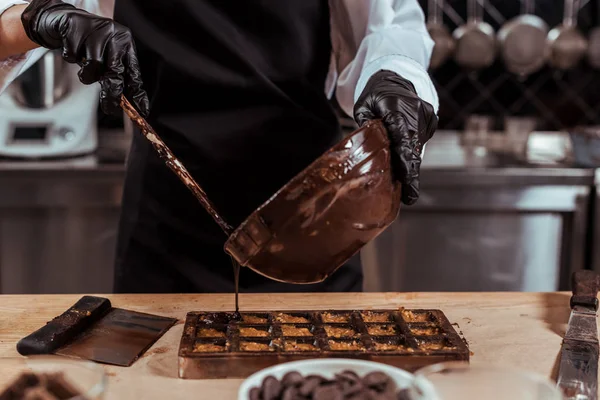 Vista recortada del recipiente de celebración chocolatero mientras se vierte chocolate derretido en moldes de chocolate — Stock Photo