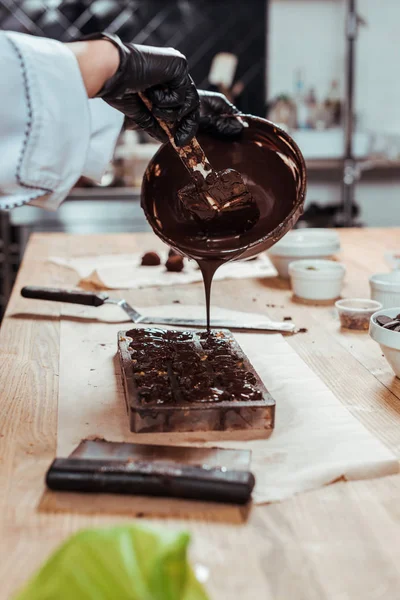 Vista ritagliata di cioccolatiera tenendo ciotola mentre versando cioccolato fuso in stampi di cioccolato sul tavolo — Foto stock