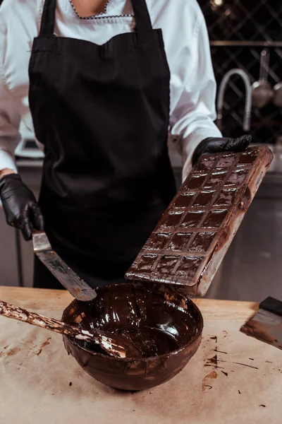 Vista recortada de chocolatero en delantal con raspador de pastel cerca de moldes de chocolate y tazón con chocolate negro derretido - foto de stock