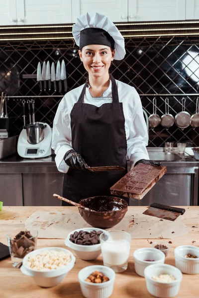 Chocolatero alegre en delantal con raspador de pastel cerca de moldes de chocolate y tazón con chocolate negro derretido - foto de stock