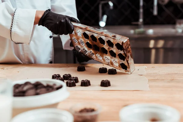 Vue recadrée du chocolatier tenant des moules au chocolat près du papier cuisson et des bonbons au chocolat — Photo de stock
