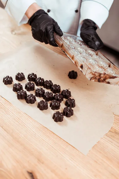 Vue recadrée du chocolatier tenant un plateau de glace près du papier cuisson et des bonbons au chocolat préparés — Photo de stock