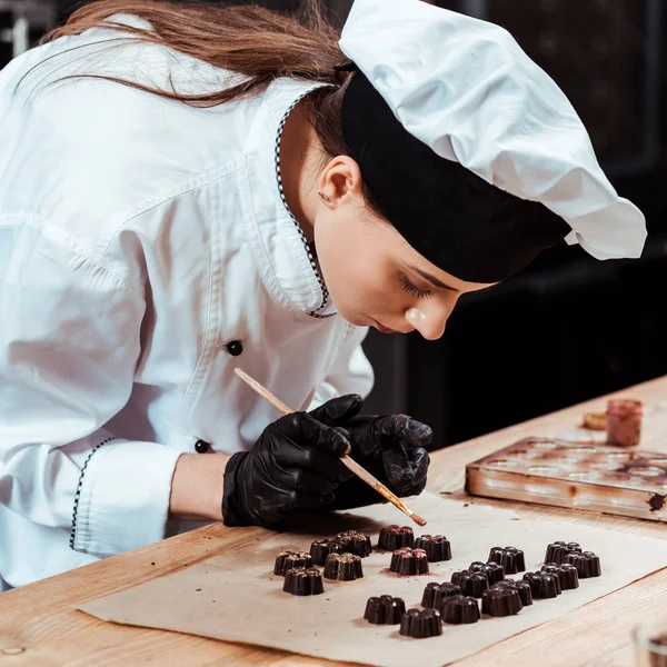 Atraente Chocolatier segurando escova com pó de ouro perto de doces de chocolate preparados no papel manteiga — Fotografia de Stock