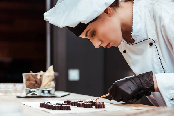 Attraktive Chocolatier Pinsel mit Goldpuder in der Nähe von leckeren Schokoladenbonbons auf Backpapier — Stockfoto
