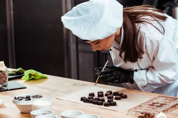 Belo chocolatier em chapéu de chef segurando escova com pó de ouro perto de doces de chocolate saborosos no papel manteiga — Fotografia de Stock