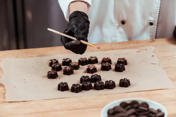Обрізаний вид шоколатьє, що тримає пензлик із золотим порошком біля смачних шоколадних цукерок на папері для випічки — стокове фото