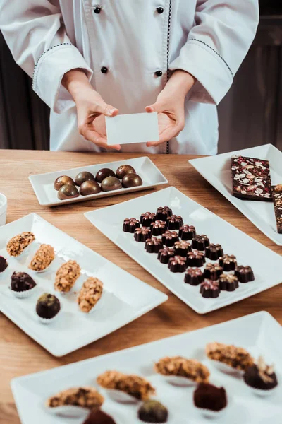Vista cortada de Chocolatier segurando cartão em branco perto de doces de chocolate saborosos em placas — Fotografia de Stock