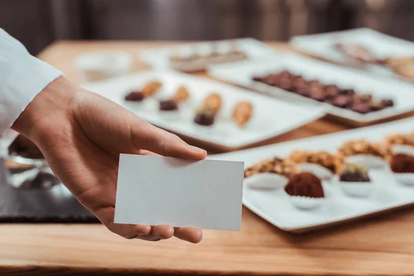 Vue recadrée du chocolatier tenant une carte blanche près de savoureux bonbons au chocolat — Photo de stock