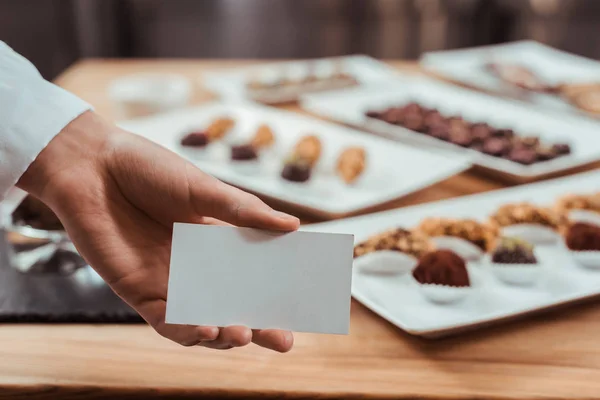 Vista ritagliata di cioccolatiere in possesso di carta bianca vicino gustose caramelle al cioccolato — Foto stock