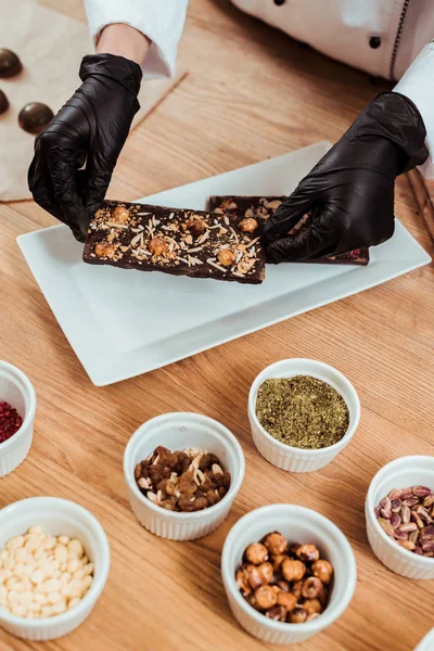 Cropped view of chocolatier in latex gloves putting chocolate bar with hazelnuts on plate — Stock Photo