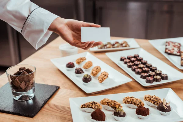 Vista cortada de Chocolatier segurando cartão em branco na mão perto de doces de chocolate saborosos em placas — Fotografia de Stock