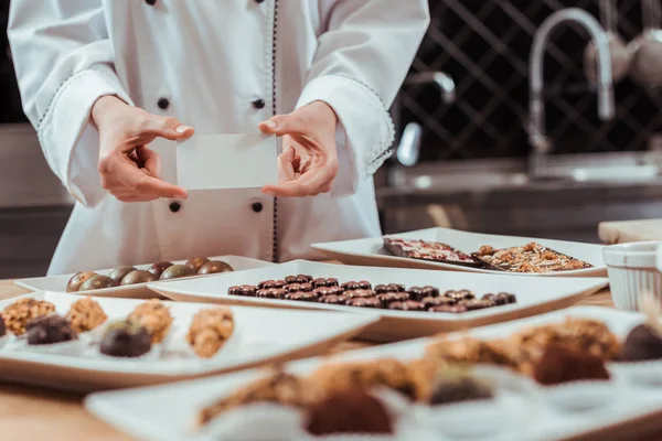 Fuoco selettivo di cioccolatiere che tengono la carta bianca in mani vicino a caramelle di cioccolato saporite su piatti — Foto stock