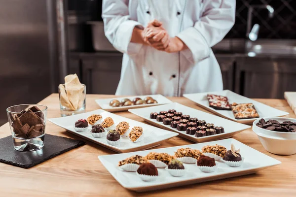 Vista recortada de chocolatero de pie con las manos cerradas cerca de sabrosos caramelos de chocolate en los platos — Stock Photo