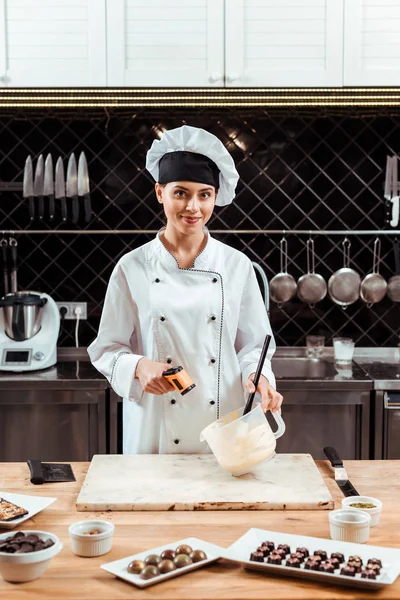 Heureux chocolatier tenant thermomètre de cuisson près du bol avec du chocolat blanc fondu — Photo de stock