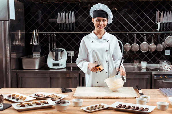 Щасливий шоколатьє в капелюсі шеф-кухаря тримає термометр для приготування їжі біля миски з розтопленим білим шоколадом — стокове фото