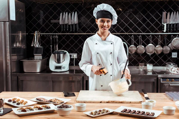 Felice cioccolatiere in cappello da chef che tiene il termometro di cottura vicino alla ciotola con cioccolato bianco fuso — Foto stock