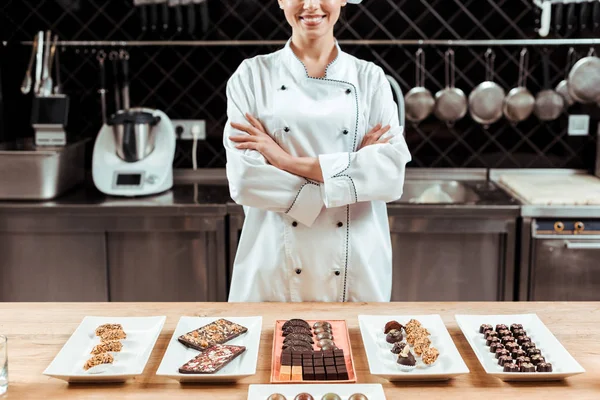 Обрізаний вид щасливого шоколатьє, що стоїть з схрещеними руками біля смачних шоколадних цукерок на тарілках — стокове фото