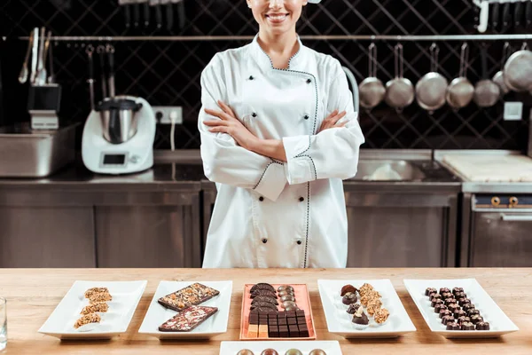 Vue recadrée de chocolatier heureux debout avec les bras croisés près de bonbons au chocolat savoureux sur des assiettes — Photo de stock