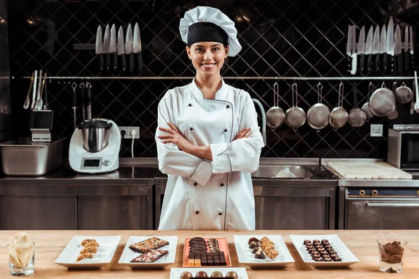 Felice cioccolatiere in cappello da chef in piedi con le braccia incrociate vicino gustose caramelle al cioccolato su piatti — Foto stock