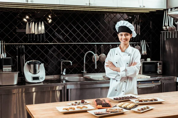 Cheerful chocolatier in chef hat standing with crossed arms near tasty chocolate candies on plates — Stock Photo