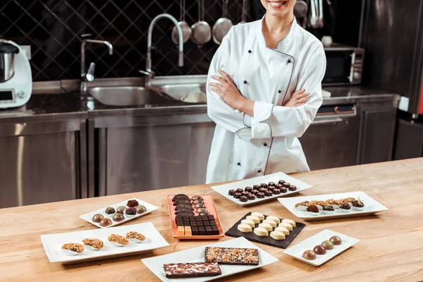 Vista cortada de chocolatier alegre de pé com braços cruzados perto de doces de chocolate saborosos em placas — Fotografia de Stock