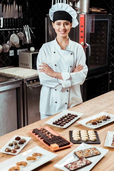 Chocolatier souriant debout avec les bras croisés près de bonbons au chocolat savoureux sur des plaques — Photo de stock