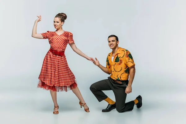 Hombre guapo de pie sobre la rodilla e invitando a la chica bonita a bailar boogie-woogie sobre fondo gris - foto de stock