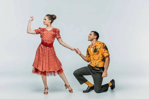 Hombre guapo de pie sobre la rodilla e invitando a la mujer bonita a bailar boogie-woogie sobre fondo gris - foto de stock