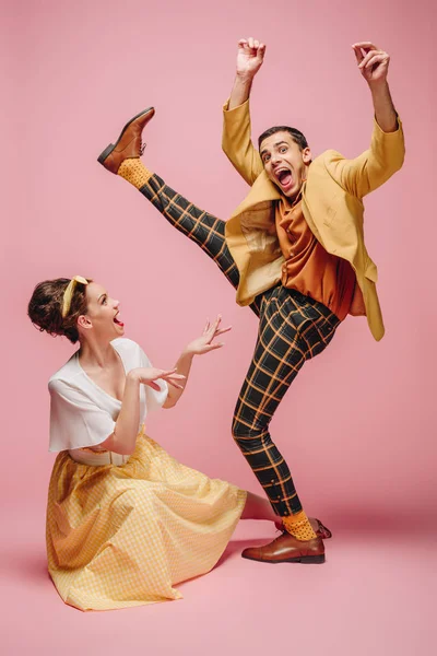 Alegre chica sentado en piso y excitado hombre levantando la pierna mientras bailando boogie-woogie sobre rosa fondo - foto de stock