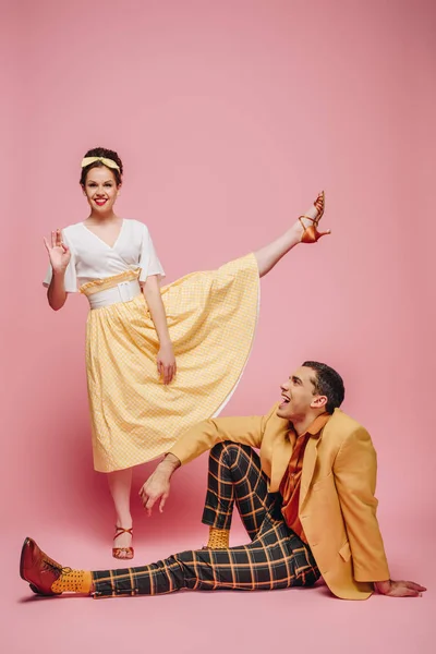 Cheerful man sitting on floor and girl waving at camera while dancing boogie-woogie on pink background — Stock Photo