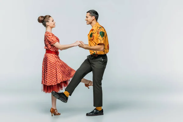 Vista laterale della coppia sorridente che si tiene per mano mentre balla boogie-woogie su sfondo grigio — Foto stock