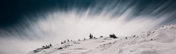 Vista panorámica de la montaña cubierta de nieve y pinos contra el cielo oscuro en la noche con nubes blancas, plano panorámico - foto de stock