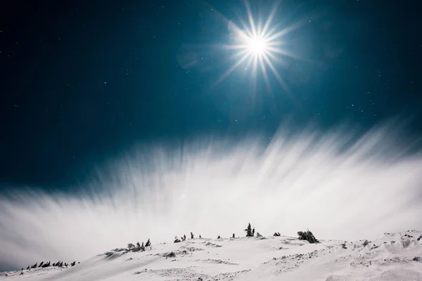 Vue panoramique sur la montagne couverte de neige et de pins contre un ciel sombre avec nuage blanc et soleil — Photo de stock