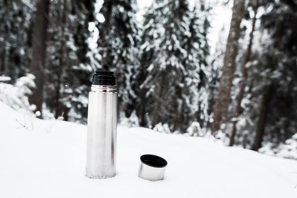 Vacuum flask with hot drink on hill covered with snow near pine forest — Stock Photo