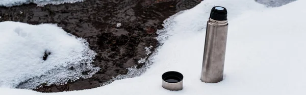 Vacuum flask on snow near flowing mountain stream, panoramic shot — Stock Photo