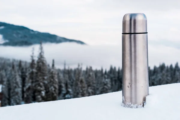 Metallic vacuum flask in snowy mountains with pine trees and white fluffy clouds — Stock Photo