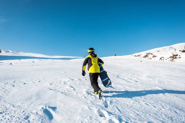 Vue arrière du sportif tenant le snowboard en marchant sur la neige — Photo de stock