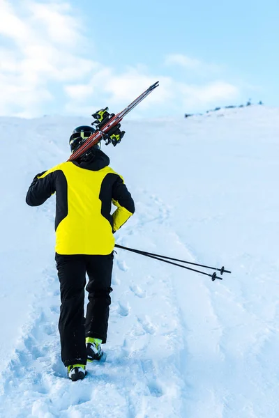 Vista trasera del esquiador en casco caminando con bastones de esquí en la nieve - foto de stock