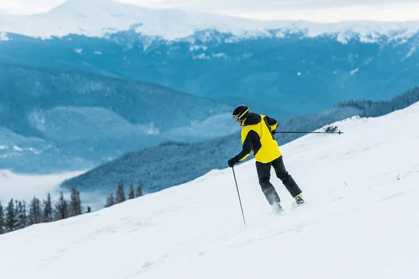 Vista trasera del esquiador sosteniendo palos y esquiando en la pista con nieve en las montañas - foto de stock