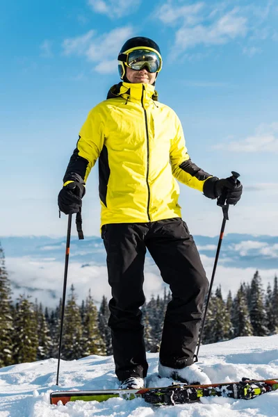 Felice sciatore in maschera guardando la macchina fotografica mentre tiene bastoni da sci contro il cielo blu — Foto stock