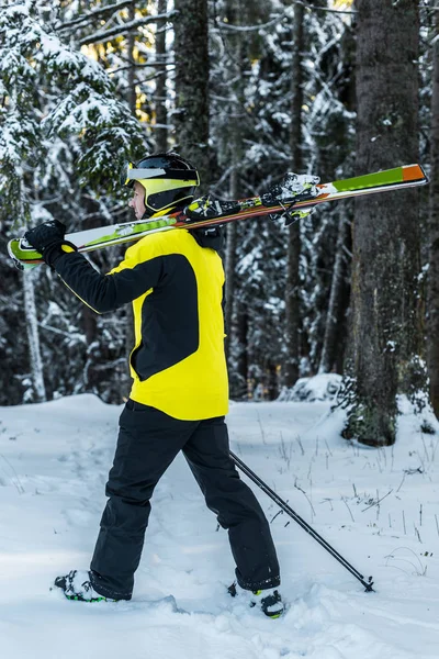 Sportsman in goggles holding ski sticks and skis while walking near firs — Stock Photo