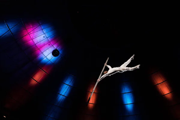 Low angle view of athletic acrobat balancing on metallic pole near blue and red lighting in arena — Stock Photo