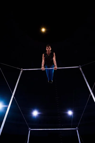 Low angle view of strong gymnast performing on horizontal bars in arena of circus — Stock Photo