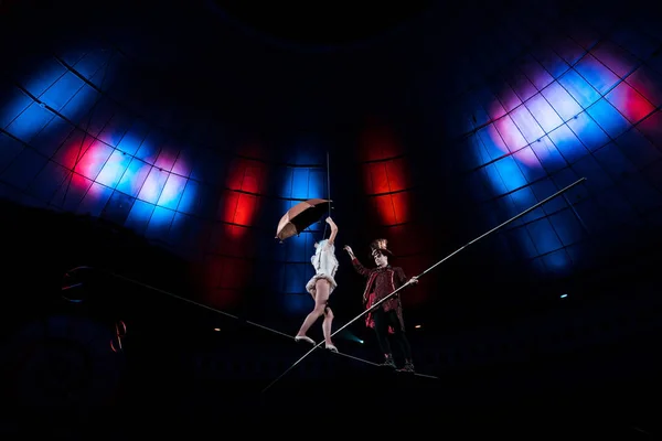 Vue à angle bas du bel homme tenant un poteau près d'un acrobate marchant avec un parapluie sur une corde — Photo de stock