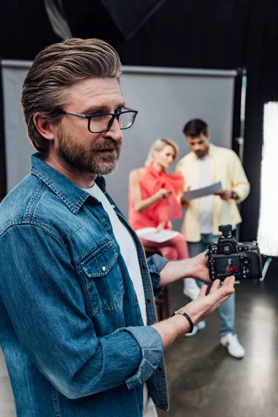 Enfoque selectivo del director de arte barbudo en gafas que apuntan con la mano a la cámara digital - foto de stock