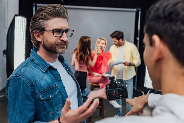 Enfoque selectivo del director de arte barbudo en gafas mirando al asistente apuntando con el dedo a la cámara digital - foto de stock