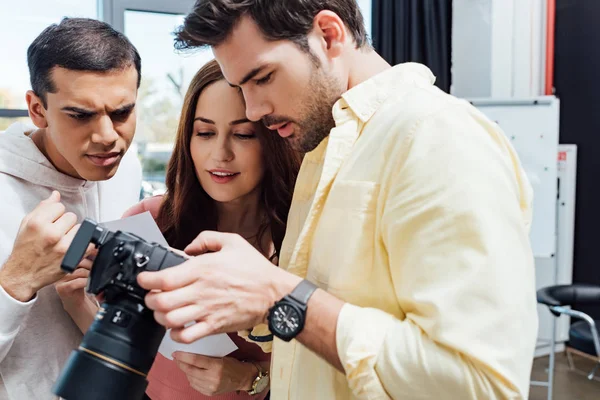 Handsome photographer looking at digital camera near coworkers — Stock Photo