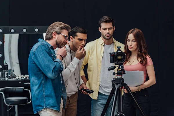 Pensativo director de arte mirando a la cámara digital en el trípode cerca de compañeros de trabajo - foto de stock