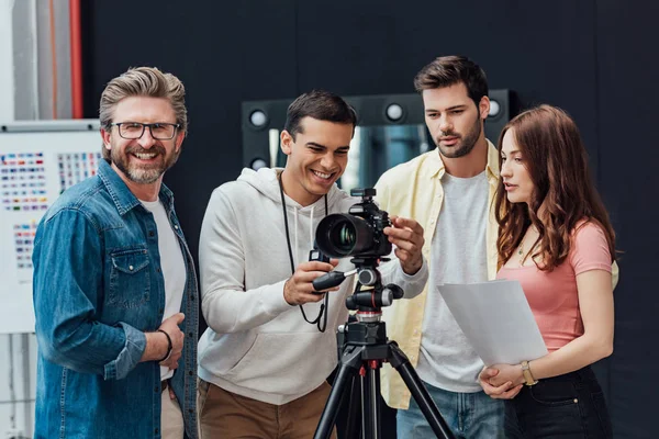 Feliz director de arte sonriendo cerca de compañeros de trabajo y cámara digital en el estudio de fotos - foto de stock